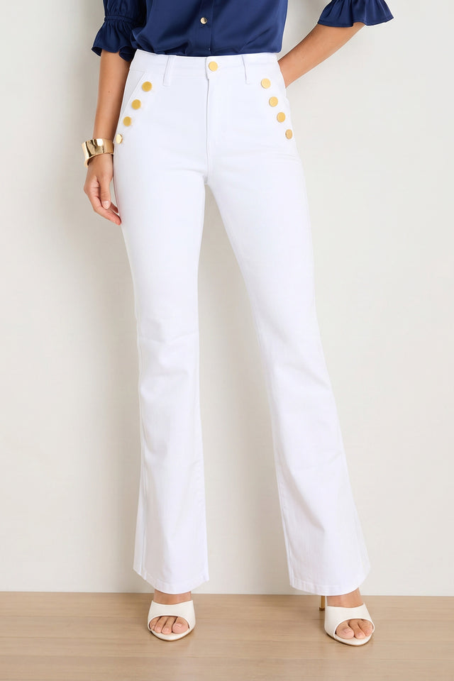Wearing the Allegra Bootcut Jean in White—a chic, high-waisted stretch style with a flared silhouette and gold buttons—a woman pairs them with a navy blouse, white heeled sandals, and a gold bracelet, standing against a light background.