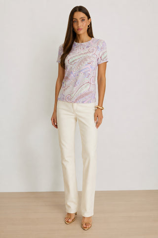 A woman stands on a wooden floor wearing the Amaya Ultra-Soft T-Shirt in Pastel Paisley with short sleeves, cream straight-leg pants, tan heeled sandals, and a gold bracelet, posing against a plain light background.