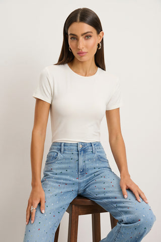 A woman with long brown hair wears the Amaya Ultra-Soft T-Shirt in white and high-waisted, light blue jeans with small colored dots as she sits on a wooden stool against a plain, light background.