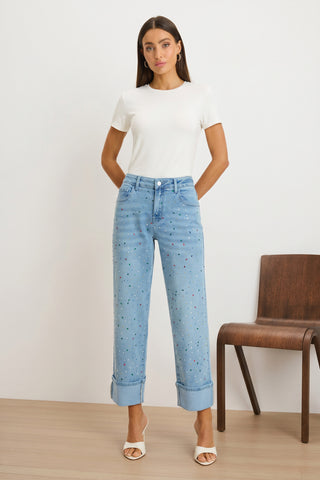 A woman stands indoors wearing the Amaya Ultra-Soft T-Shirt in white with high-waisted embellished light blue jeans and white heeled sandals, hands behind her back, next to a wooden chair.