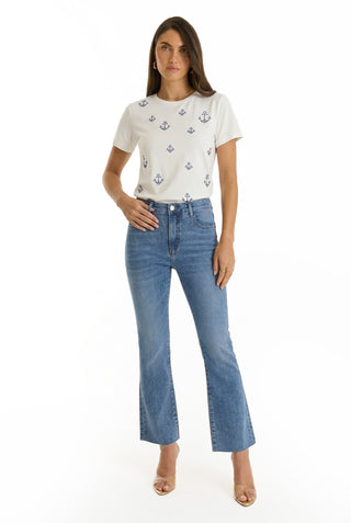 A woman stands against a white background wearing a white t-shirt with blue anchors, Nicolette Cropped Jean - Medium Blue in four-way stretch denim, and clear high-heeled shoes for effortless summer style.