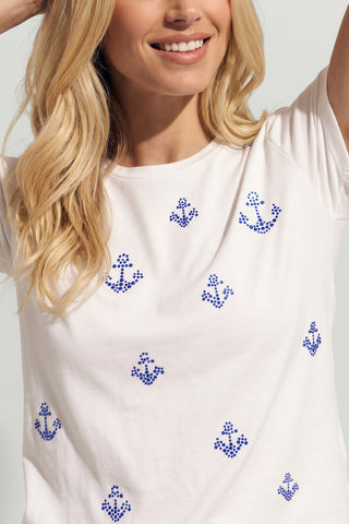 A woman with long blonde hair smiles and raises her arm, wearing the Amaya Ultra Soft T-Shirt - Crystal Anchors, featuring small blue anchor prints for a nautical-inspired look.