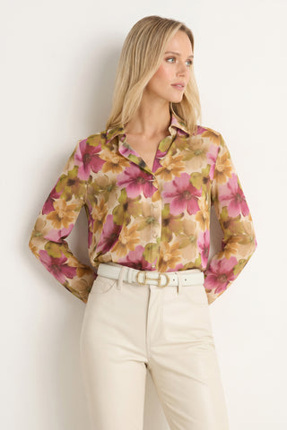 A woman with long blonde hair stands against a white wall, looking to her left while effortlessly showcasing the Ana Chiffon Blouse - Floral Print, featuring vibrant pink, yellow, and green flowers. She pairs the versatile women’s top with high-waisted cream pants and a white belt.