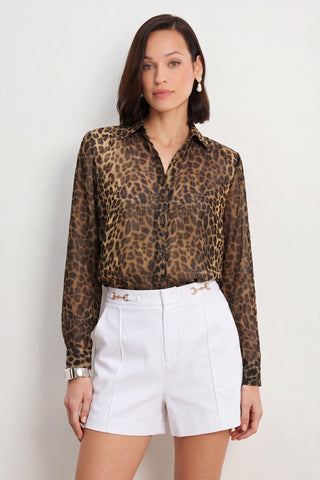 Wearing the Ana Chiffon Blouse - Leopard with long sleeves, she pairs it with high-waisted white shorts featuring gold chain details, accessorized with earrings and a bracelet, standing against a plain light background.