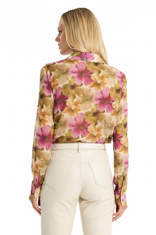 A woman with long blonde hair is seen from the back, wearing the Ana Chiffon Blouse in a floral print, featuring pink, yellow, and green hues. She pairs it with high-waisted off-white pants against a plain white background.
