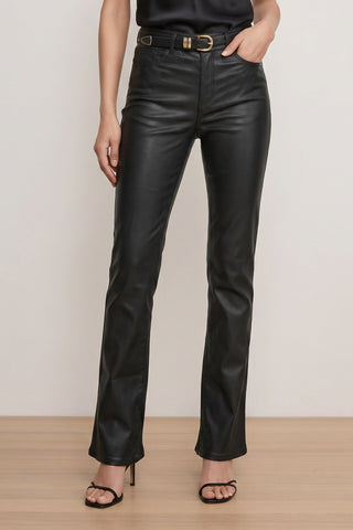 Wearing the Aria Coated Flare Jean - Black, styled with a black belt and black high-heeled sandals, a person stands on a wooden floor against a light wall. Only the lower half is visible; the upper body and face are not shown.