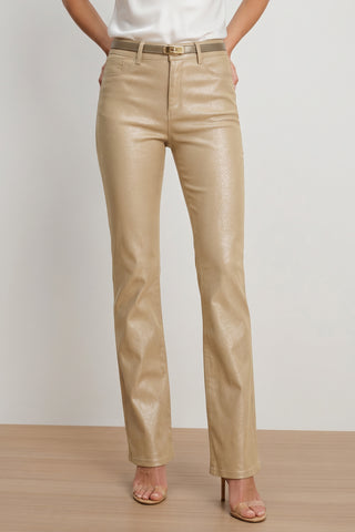 A person wearing Aria Coated Flare Jean in Champagne with a matching belt, a white top, and beige heeled sandals, standing on a wooden floor against a plain light background.
