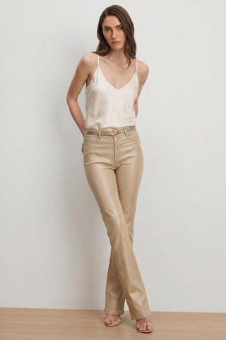 A woman with long brown hair and a neutral expression stands against a plain wall wearing the Aria Coated Flare Jean in Champagne, a cream satin camisole, and open-toe heeled sandals.