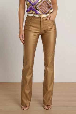 A person stands indoors in Aria Coated Flare Jean - Gold Metallic, a high-waisted, statement piece with a metallic finish and flared silhouette. Paired with a white belt, patterned sleeveless top, open-toe heels; plain neutral background.