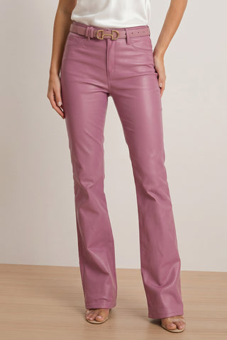 A woman stands on a wooden floor against a plain background, wearing the Aria Coated Flare Jean in mauve with a matching belt, styled with a white top and open-toe nude heels.