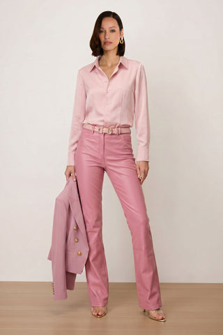 A woman stands against a plain backdrop wearing the Aria Coated Flare Jean in Paradise Pink, paired with a matching pink button-up shirt, a pink belt, open-toe heels, and holding a pink blazer.