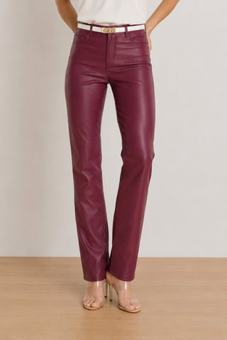 Wearing the Aria Coated Flare Jean in Sangria—a high-waisted pair with a subtle coated finish—styled with a white belt and clear-heeled sandals, this person stands on a light wood floor against a plain light background.