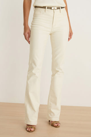 A person wearing Aria Coated Flare Jean in Vanilla, styled with a beige belt and brown open-toe heels, standing on a light wooden floor against a plain white background.