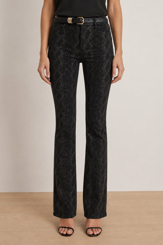 A person wearing the Aria Flare Jean - Black Python, featuring a striking snakeskin-patterned design, paired with a black belt with a gold buckle and black open-toe heels, stands against a neutral background. The image is cropped to show only the lower body.
