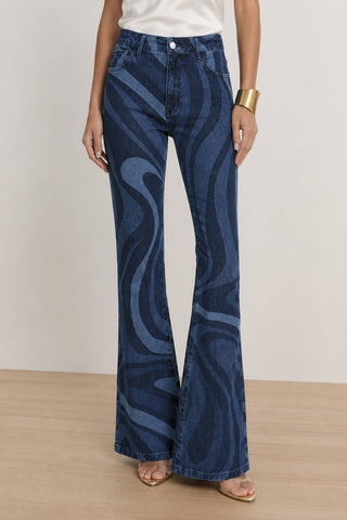A person wearing the Aria Flare Jean - Blue Wave with a wavy pattern, paired with a white top, gold bracelet, and open-toe sandals stands on a wooden floor against a light background.