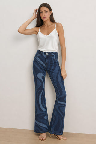 A woman with long brown hair and a neutral expression stands indoors against a plain wall, wearing the Aria Flare Jean - Blue Wave, a white satin camisole, and beige heeled sandals.