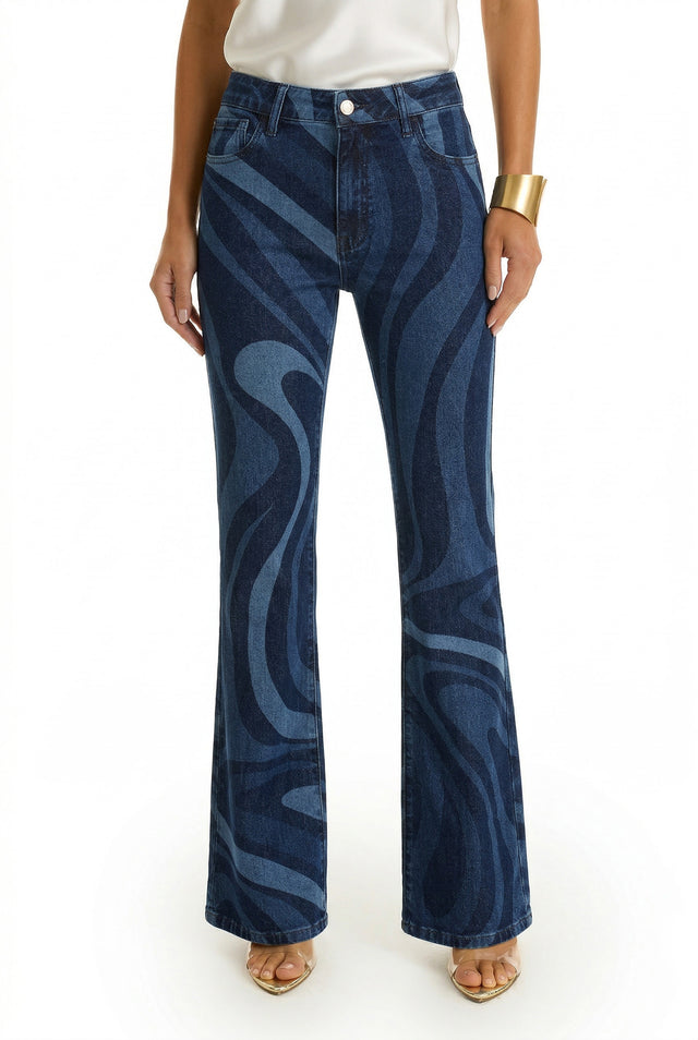 A person wearing the Aria Flare Jean - Blue Wave, featuring a wavy abstract pattern, with a white top, gold cuff bracelet, and open-toe heels. The image is cropped from shoulders down on a white background.