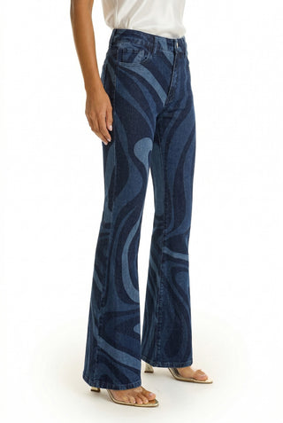 Someone wears the Aria Flare Jean - Blue Wave, featuring a wavy abstract blue design, styled with a white sleeveless top and open-toed heeled sandals. Only the lower half of their body is shown.