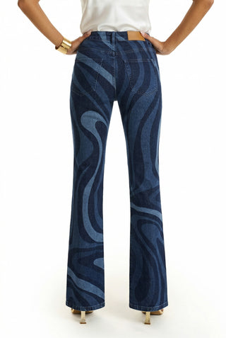 A woman stands with hands on hips, wearing the Aria Flare Jean - Blue Wave and a white top. Seen from behind against a plain white background, she showcases the stylish blue wave-patterned bootcut denim.