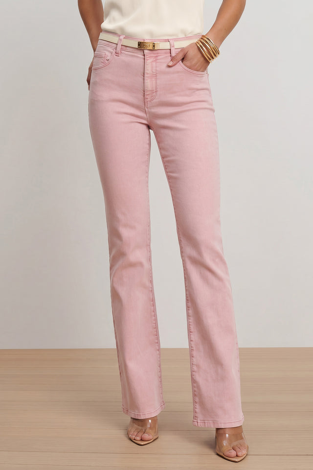 Wearing the Aria Flare Jean - Blush, a cream top, gold bangles, and transparent heeled sandals, a person stands with one hand in their pocket against a neutral background.