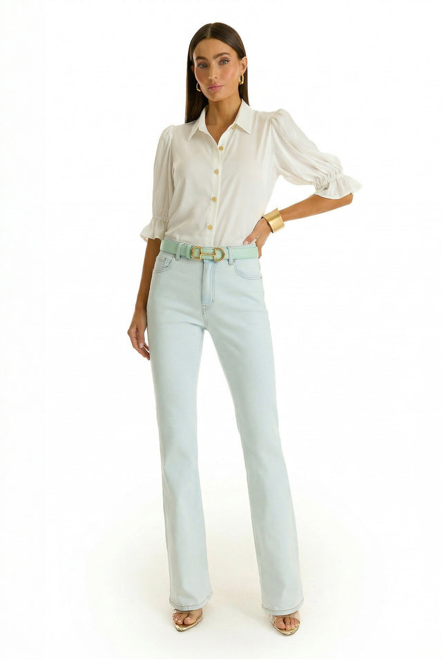 A woman with straight brown hair stands against a white background wearing the Aria Flare Jean - Cloud, a white button-up blouse with puffed sleeves, a beige belt, gold bangle, and open-toe heels. She has a neutral expression.