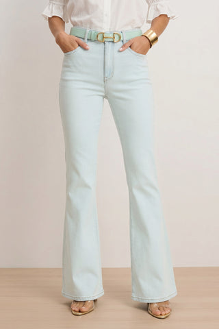 Wearing the Aria Flare Jean - Cloud with a matching belt, a white blouse, gold bangle, and clear heeled sandals, a person stands hands-in-pockets against a light background.