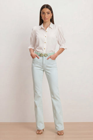 A woman with long straight hair stands against a plain background wearing the Aria Flare Jean - Cloud, a white puff-sleeve blouse, a mint green belt, and tan heeled sandals, with her hands in her pockets.