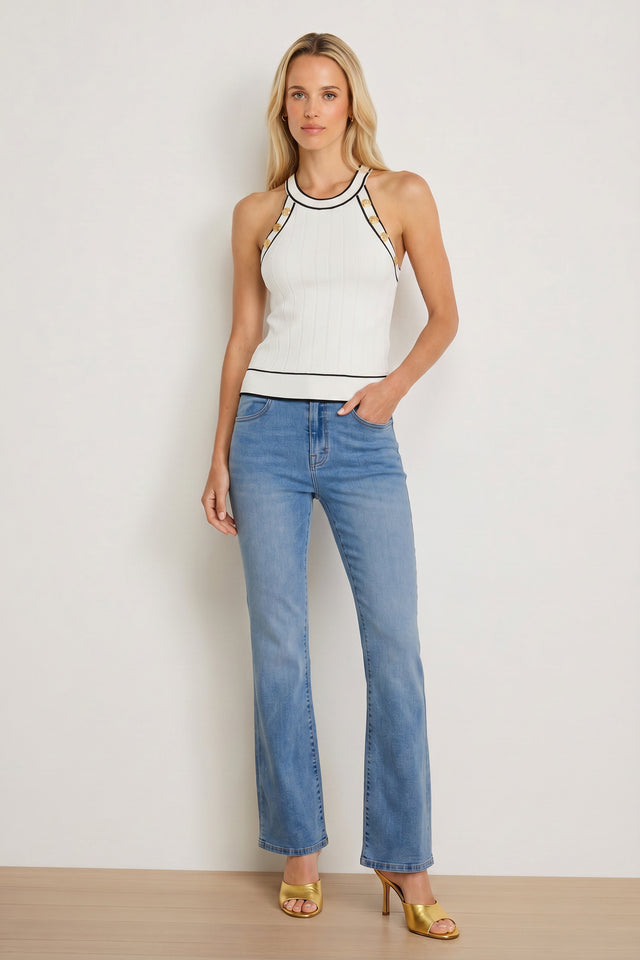 A woman faces the camera with a neutral expression, one hand in her pocket, wearing a white sleeveless top with black trim, gold high-heeled sandals, and the Aria Flare Jean - Medium Blue against a plain wall.