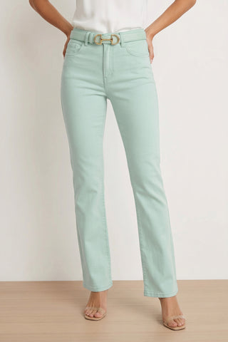 A person wears the Aria Flare Jean in mint with a matching belt, a white top, and clear open-toe heels—standing on a light wooden floor against a white wall, highlighting the jean's soft Four-Way Stretch Denim.