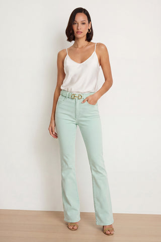A woman stands against a white wall, wearing a white camisole, Aria Flare Jean - Mint with Four-Way Stretch Denim and matching belt, tan heeled sandals, and a side-parted bob hairstyle.