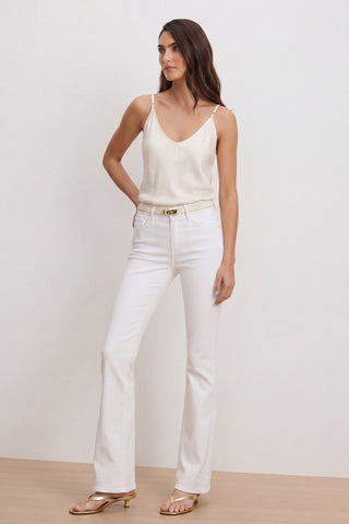 A woman poses confidently in a cream camisole, gold heeled sandals, and the Aria Flare Jean - White, featuring high-rise style and four-way stretch. She has long wavy brown hair and stands against a plain background.