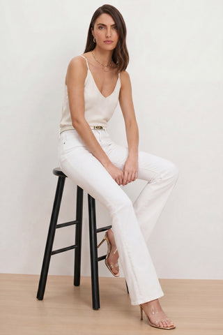A woman with long brown hair sits on a black stool against a plain light background, wearing a cream camisole, a belt, clear high-heeled sandals, and the Aria Flare Jean - White featuring four-way stretch and high-rise fit.