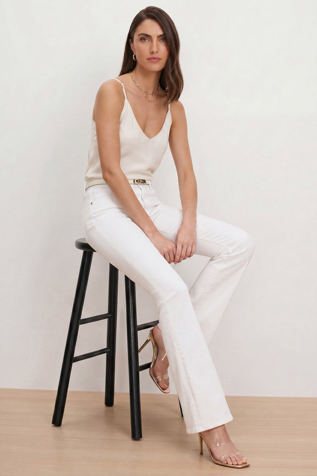 A woman with long brown hair sits on a black stool against a plain light background, wearing a cream camisole, a belt, clear high-heeled sandals, and the Aria Flare Jean - White featuring four-way stretch and high-rise fit.