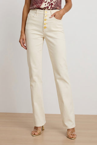 A person wearing the Aubrey Bootcut Jean in ivory, paired with a sleeveless patterned top and transparent heeled sandals, stands on a wooden floor against a plain background.