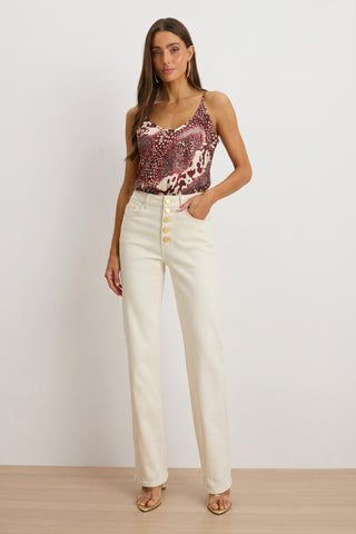Woman stands against a plain background wearing a red and white sleeveless top, the Aubrey Bootcut Jean - Ivory with large buttons, gold open-toe heels, and has one hand casually tucked in her pocket.