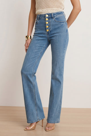 Wearing Aubrey Bootcut Jean - Medium Blue with gold buttons, a sleeveless crochet top, clear high-heeled sandals, and a gold cuff bracelet, a person stands on a wooden floor against a light backdrop.
