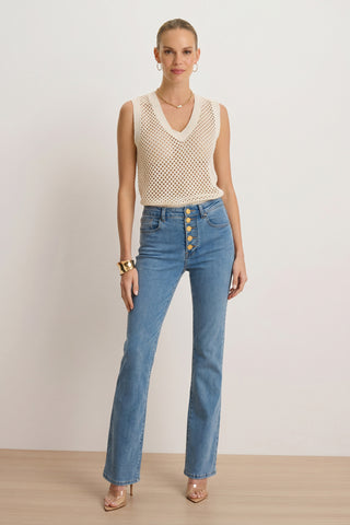 A woman faces forward, smiling slightly, in a sleeveless cream knit top, gold jewelry, clear-heeled shoes, and the Aubrey Bootcut Jean - Medium Blue—made with four-way stretch denim for comfort.
