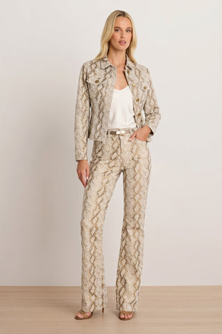A woman with long blonde hair stands indoors against a plain light wall, confidently wearing the Bethanie Denim Jacket - Gold Python, matching pants, a white camisole, nude heels, and minimal jewelry.