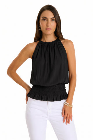 A woman with long brown hair models the Betsy Halter Top in black, featuring a sleeveless, ruched waist design, paired with white pants. She poses with one hand on her hip against a plain white background.