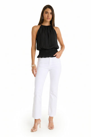 A woman stands against a white background wearing the Betsy Halter Top - Black, paired with white high-waisted pants, clear heeled sandals, and wrist jewelry. This chic halter top adds an elegant touch to her modern style.