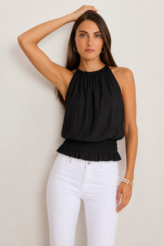 A woman stands against a neutral background, wearing the Betsy Halter Top in black—a perfect piece for summer fashion—paired with high-waisted white jeans, with one hand resting on her head and the other at her side.