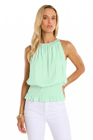 A woman with long blonde hair models an elevated resort look: the sleeveless Betsy Halter Top - Mint paired with white pants and gold bracelets. She poses in front of a plain white background, one arm on her hip.
