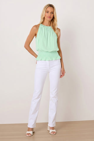 A woman stands against a plain background in the Betsy Halter Top - Mint, a feminine light green summer top with a gathered waist, paired with white pants and heeled sandals—the perfect vacation look.
