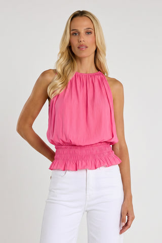 A woman with long blonde hair wears the Betsy Halter Top - Petal Pink, a feminine summer top featuring a smocked waist and ruffled hem, styled with high-waisted white pants against a plain light background for an effortless vacation look.