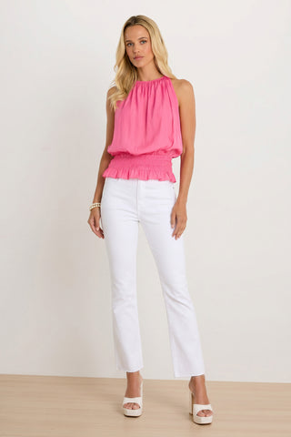 A woman with long wavy blonde hair stands on a wooden floor in front of a light background, wearing the Betsy Halter Top - Petal Pink, styled with white high-waisted pants, platform heels, and a bracelet for a summery vacation look.