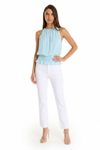 A woman with straight brown hair poses confidently against a white background, wearing the Betsy Halter Top - Sky Blue styled with white high-waisted pants and nude open-toe heels.