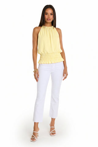 A woman stands against a white background wearing the Betsy Halter Top - Sunshine Yellow, a sleeveless, feminine top with a ruffled waist, styled with white cropped pants, heeled sandals, and gold bangles for an effortless summer look.