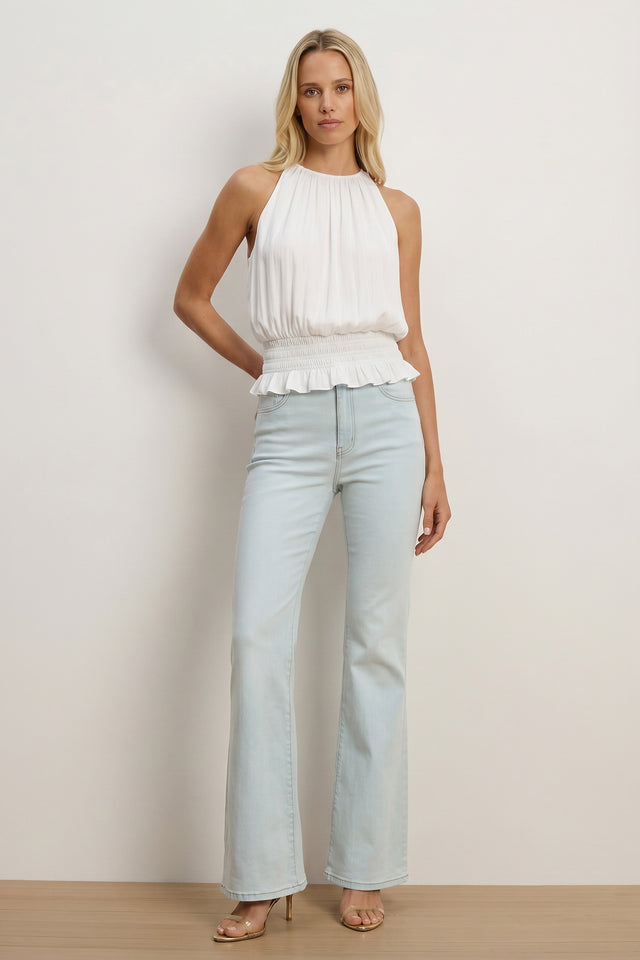 A woman poses in the Betsy Halter Top - White, featuring a feminine halter neckline and ruffled waist, styled with light blue high-waisted flared jeans and beige heels—an ideal choice for effortless summer fashion.