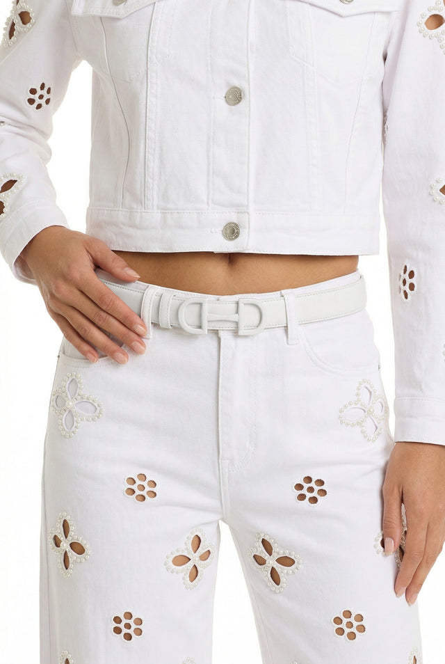 A person wears a white denim jacket and pants set with floral cut-outs, silver buttons, and embroidery, styled with the Bettie Belt - Blanc featuring a horsebit buckle. They pose with one hand on their waist.