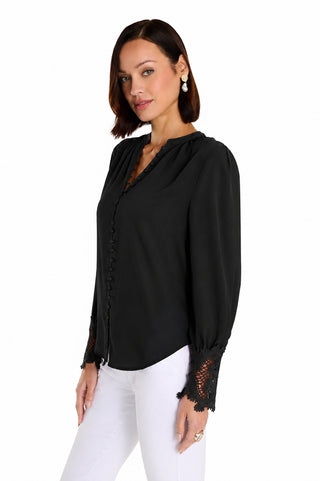 A woman with short brown hair wears the Blake Lace Sleeve Blouse in black with lace cuffs and white pants. She stands slightly turned against a white background, accessorized with pearl drop earrings.